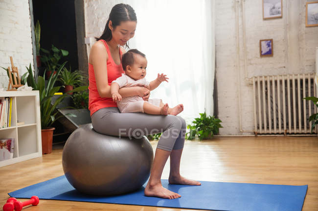Bela sorrindo jovem asiático mulher sentado com bebê no fitness bola em casa — Fotografia de Stock