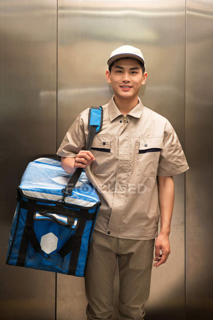 Guapo joven asiático mensajero con bolsa sonriendo a cámara en ascensor - foto de stock