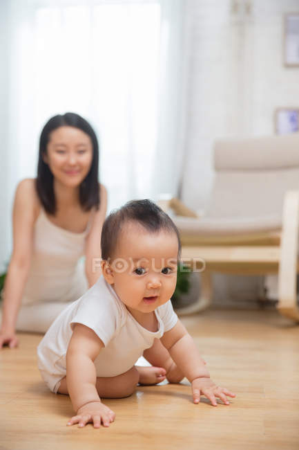 Прелестный азиатский ребенок ползает по полу и смотрит в камеру, счастливая мать сидит позади — стоковое фото