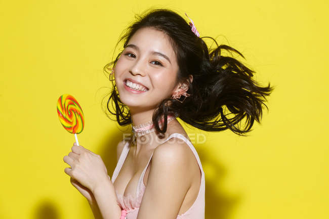 Beautiful happy young woman holding colorful lollipop and smiling at camera on yellow — Stock Photo