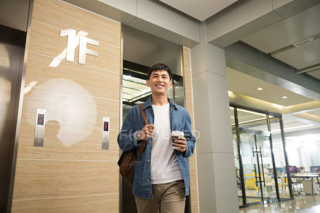 Low angle view of smiling young asian businessman holding coffee to go and walking near elevator in office — Stock Photo