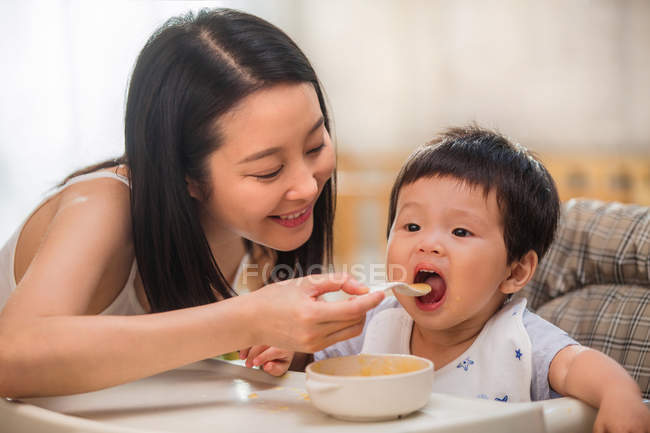 Happy young mother holding spoon and feeding cute toddler at home — Stock Photo