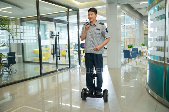 Улыбающийся молодой азиатский охранник едет на самобалансирующемся скутере и использует рацию в офисе — стоковое фото