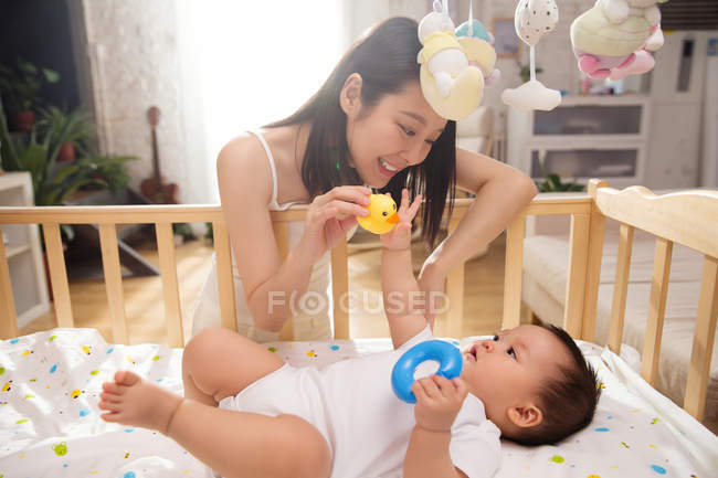 Счастливая молодая мать, держащая резиновую утку и играя с очаровательным младенцем, лежащим в кроватке — стоковое фото