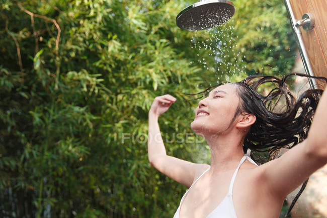 Feliz joven asiática mujer en bikini lavado de cabello y tomando ducha con los ojos cerrados verde fondo natural - foto de stock