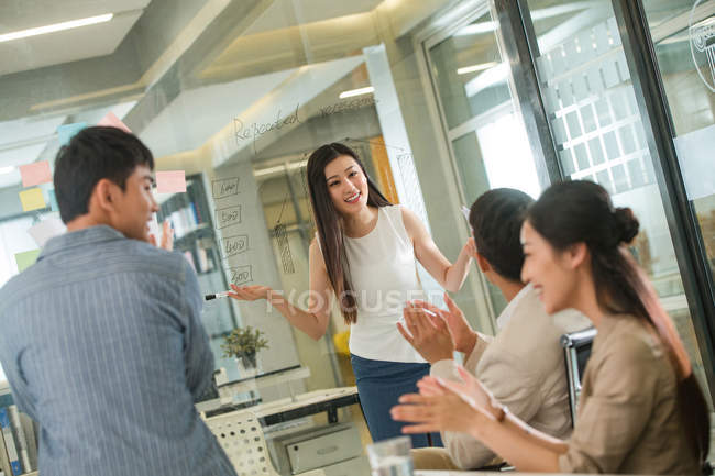 Jeunes collègues d'affaires applaudissant tout en souriant femme d'affaires parlant près du verre avec des notes dans le bureau — Photo de stock