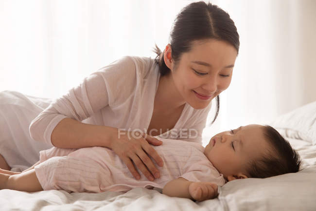 Beautiful happy young asian woman looking at her adorable baby sleeping on bed — Stock Photo
