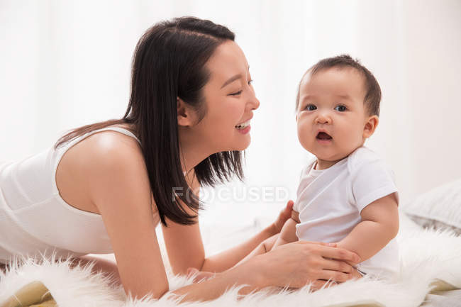 Vista laterale di sorridere giovane donna asiatica guardando carino neonato seduto su pelliccia coperta sul letto — Foto stock