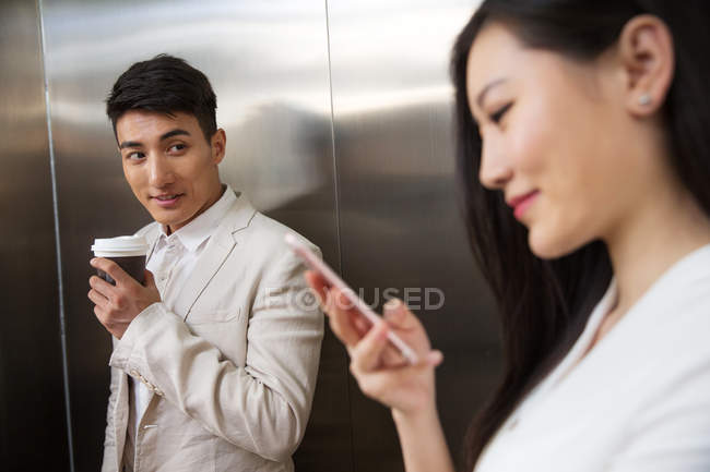 Jeune homme d'affaires tenant du café pour aller et regardant belle femme d'affaires en utilisant smartphone au premier plan dans l'ascenseur — Photo de stock