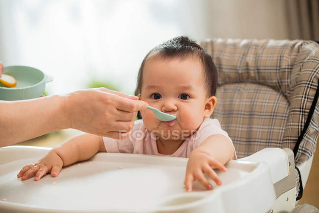 Tiro cortado de tigela mãe segurando com colher e alimentação adorável bebê em casa — Fotografia de Stock