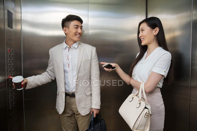 Молодой бизнесмен с кофе идти и бизнесмен со смартфоном улыбается друг другу в лифте — стоковое фото