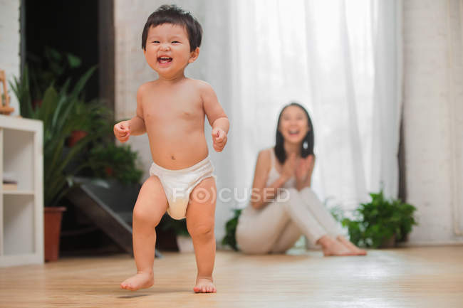 Feliz joven madre aplaudiendo de la mano y mirando a su adorable niño pequeño empezando a caminar en casa - foto de stock