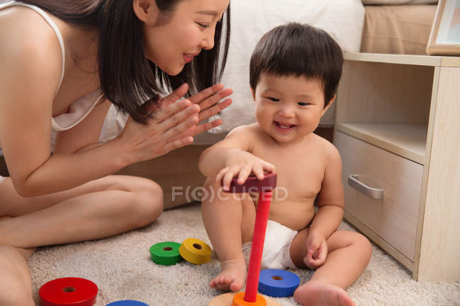 Schnappschuss einer glücklichen jungen Mutter, die in die Hände klatscht und lächelndes Baby beim Spielen mit buntem Spielzeug zu Hause ansieht — Stockfoto