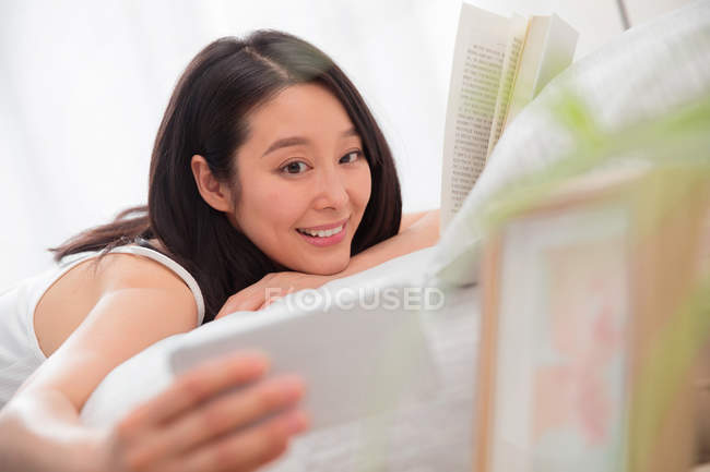 Selettiva messa a fuoco di sorridere giovane donna asiatica prendendo selfie con smartphone durante la lettura di libro sul letto — Foto stock