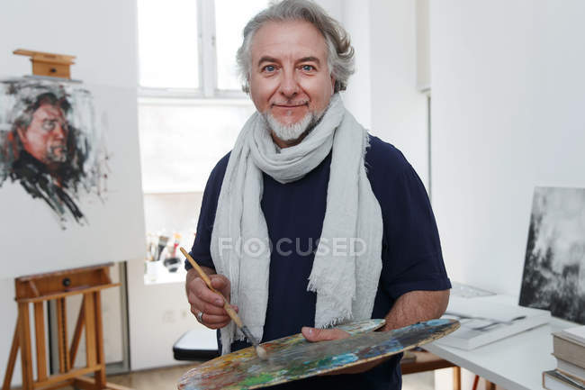 Heureux mature mâle artiste tenant palette avec pinceau et souriant à la caméra en studio — Photo de stock