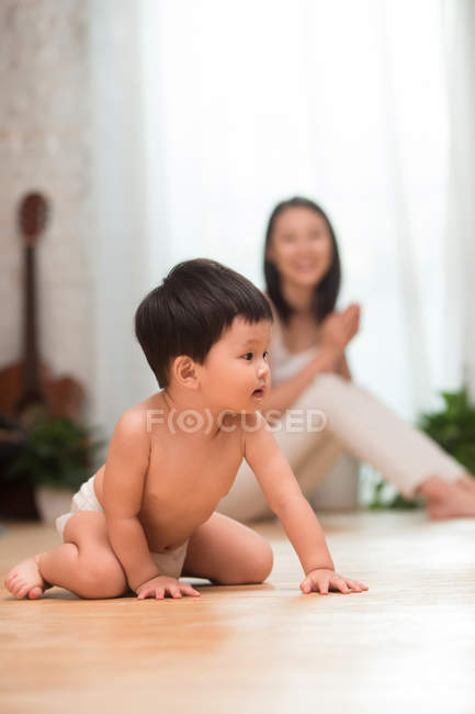 Adorável asiático criança em fralda agachamento no chão enquanto feliz mãe sentado atrás, seletivo foco — Fotografia de Stock