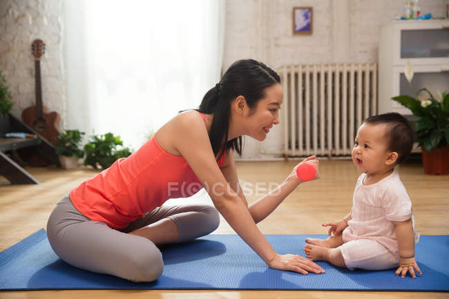 Vista lateral de la sonriente madre joven sosteniendo la mancuerna y mirando al adorable bebé sentado en la esterilla de yoga - foto de stock