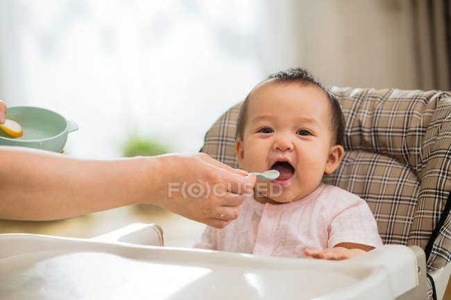 Cropped shot of mother holding spoon and feeding adorable asian baby — Stock Photo