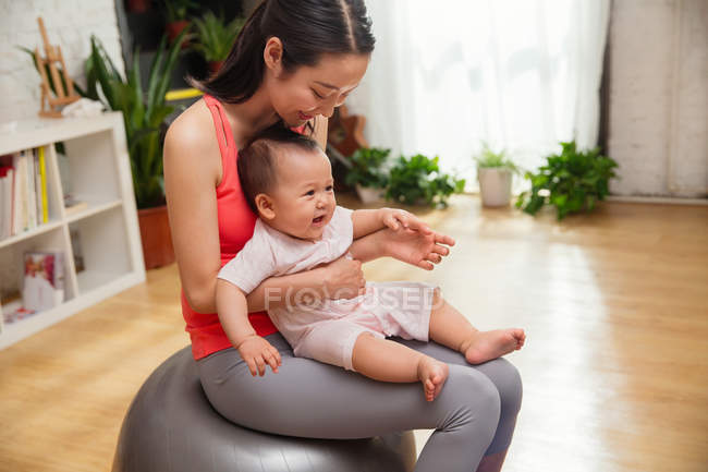 Feliz joven madre en ropa deportiva sentado en la pelota de fitness con bebé emocionado - foto de stock