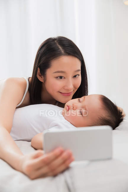 Happy young mother holding smartphone and taking selfie with baby sleeping on bed — Stock Photo