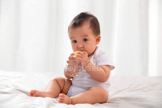 Полный вид восхитительного азиатского младенца, сидящего на кровати и питьевой воды из детской бутылочки — стоковое фото