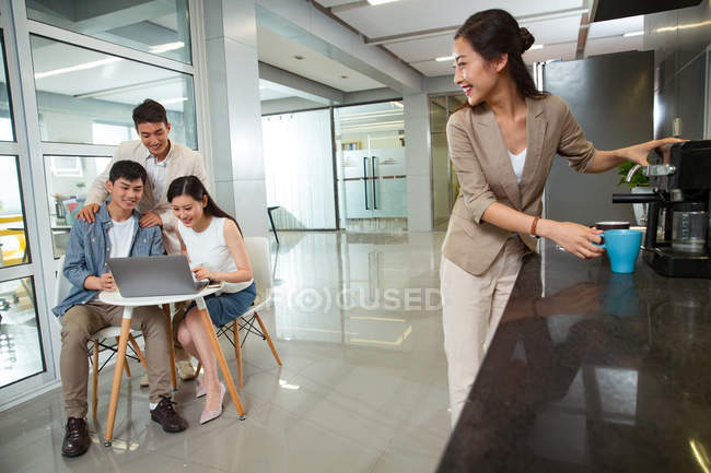 Усміхнена молода бізнес-леді робить каву і дивиться на колег, використовуючи ноутбук в офісі — стокове фото