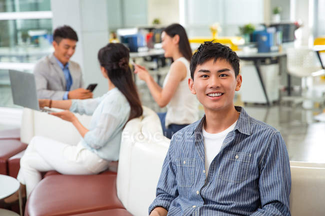 Bonito feliz jovem empresário sorrindo para câmera enquanto colegas que trabalham para trás no escritório — Fotografia de Stock