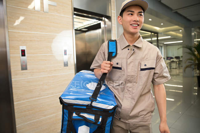 Улыбающийся молодой азиатский курьер с сумкой, смотрящий в бизнес-центр — стоковое фото