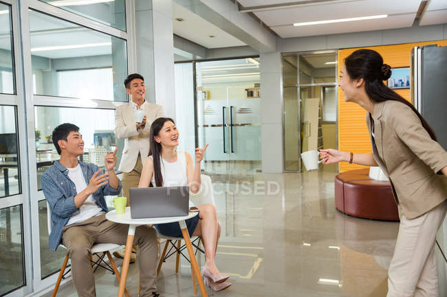 Feliz joven empresaria sosteniendo la taza de café y hablando con colegas alegres usando el ordenador portátil durante el descanso en la oficina - foto de stock