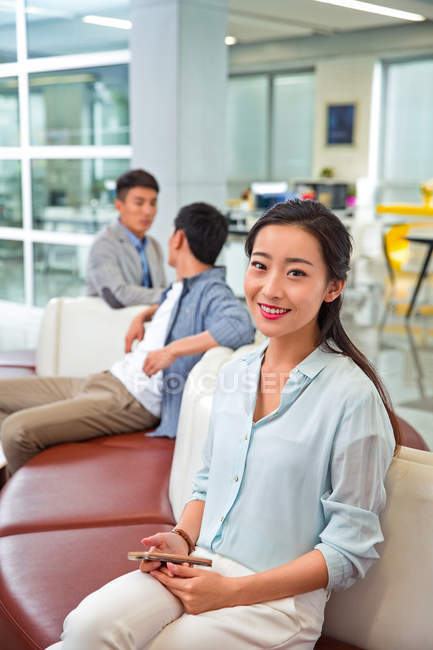 Красивая молодая деловая женщина, держащая смартфон и улыбаясь в камеру, сидя в офисе — стоковое фото