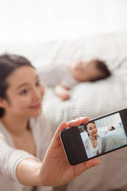 Messa a fuoco selettiva della giovane madre che prende selfie con smartphone mentre il bambino dorme sul letto — Foto stock
