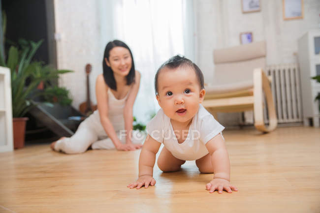 Adorabile asiatico bambino strisciare su pavimento e guardando fotocamera felice madre seduta dietro — Foto stock