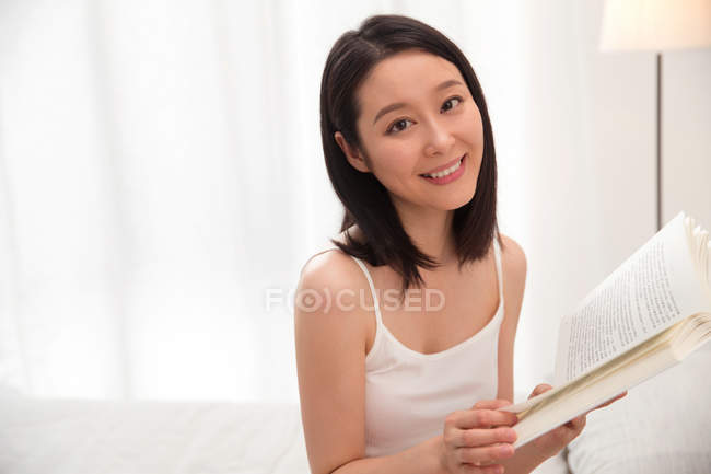 Schöne junge asiatische Frau hält Buch und lächelt in die Kamera — Stockfoto