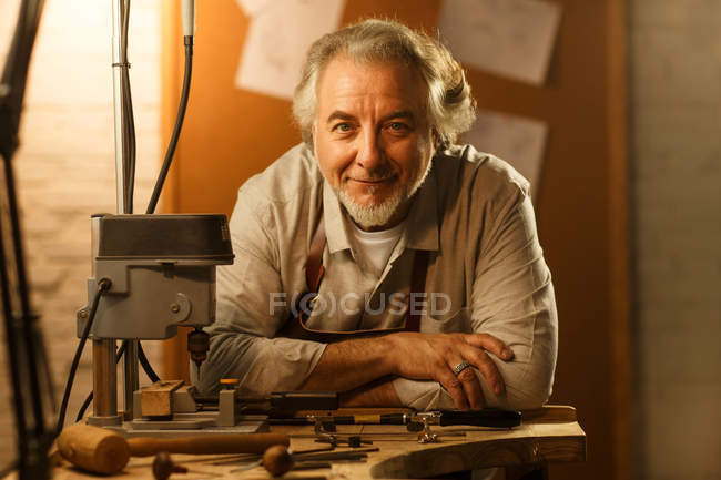 Professional jewelry designer in apron leaning at table and smiling at camera in workshop — Stock Photo