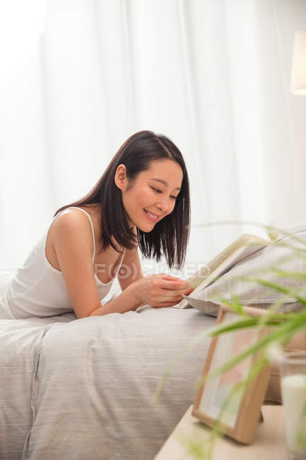 Красивая улыбающаяся молодая азиатка лежит в постели и читает книгу — стоковое фото