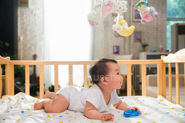 Вид збоку чарівна азіатська дитина лежить в ліжечку і дивиться на барвисті іграшки, що висять зверху — стокове фото