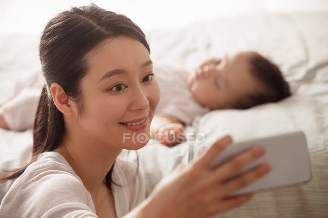 Beautiful smiling young mother taking selfie with smartphone while baby sleeping behind in bedroom — Stock Photo