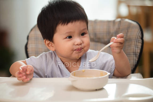 Чарівний азіатський малюк тримає ложку і дивиться геть під час їжі вдома — стокове фото