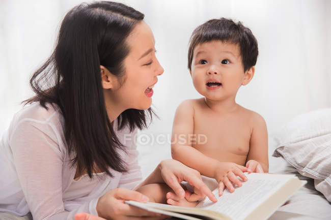Happy young mother reading book with adorable infant child sitting on bed — Stock Photo
