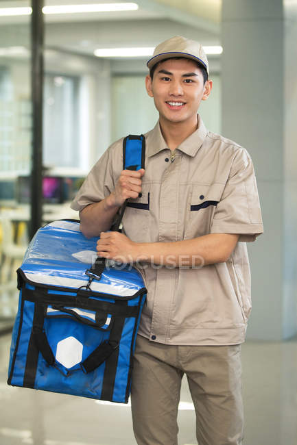 Красивый молодой азиатский курьер с сумкой, улыбающейся в камеру в офисе — стоковое фото