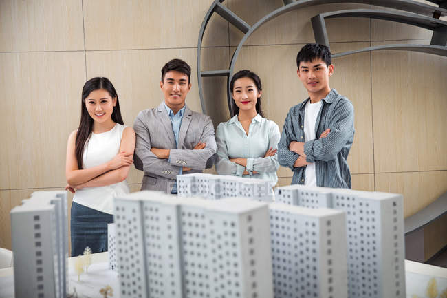 Profesionales jóvenes asiáticos arquitectos de pie con los brazos cruzados cerca de proyecto y sonriendo a la cámara en la oficina - foto de stock