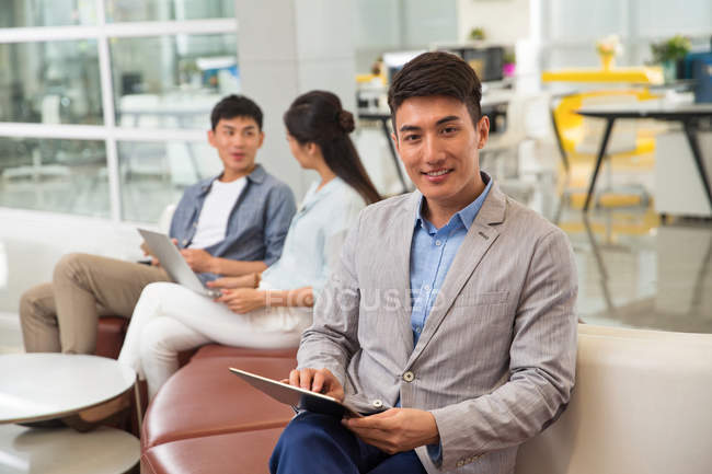 Красивый молодой бизнесмен, использующий цифровой планшет и улыбающийся на камеру в офисе — стоковое фото