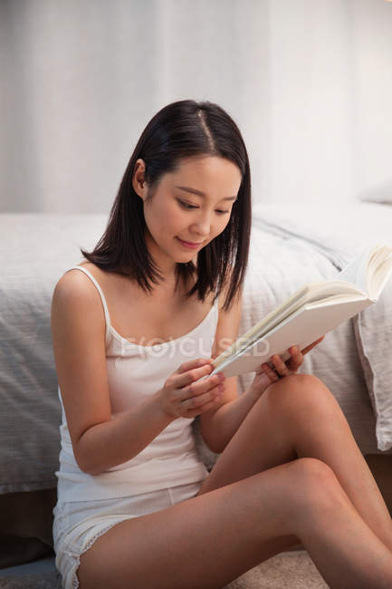 Hermosa sonriente joven asiático mujer sentado cerca de cama y lectura libro - foto de stock