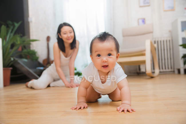Sorrindo jovem mãe olhando para o belo bebê rastejando no chão em casa — Fotografia de Stock
