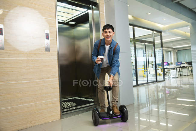 Красивий щасливий молодий азіатський бізнесмен тримає каву, щоб піти і їздити на самобалансуючому скутері біля ліфта — стокове фото