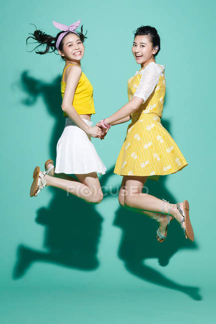 Seitenansicht glücklicher asiatischer Freundinnen, die Hände halten und auf blau springen — Stockfoto