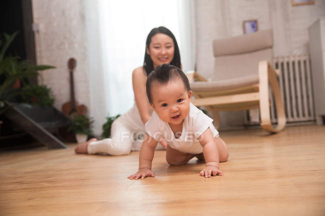 Glückliche junge Mutter blickt auf entzückendes Baby, das auf dem Boden kriecht und in die Kamera lächelt — Stockfoto