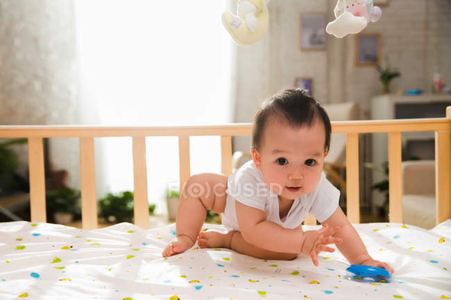 Очаровательный азиатский ребенок, держащий синюю игрушку и смотрящий в камеру во время игры в кроватке — стоковое фото