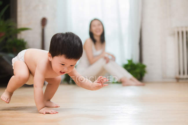 Adorable feliz asiático niño en pañal arrastrándose en suelo mientras sonriendo madre sentado detrás, enfoque selectivo - foto de stock