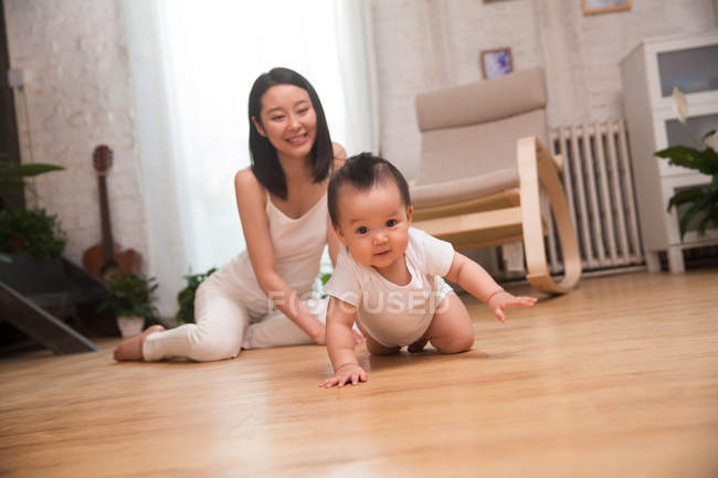Vista completa de la feliz madre joven mirando al bebé arrastrándose en el suelo - foto de stock